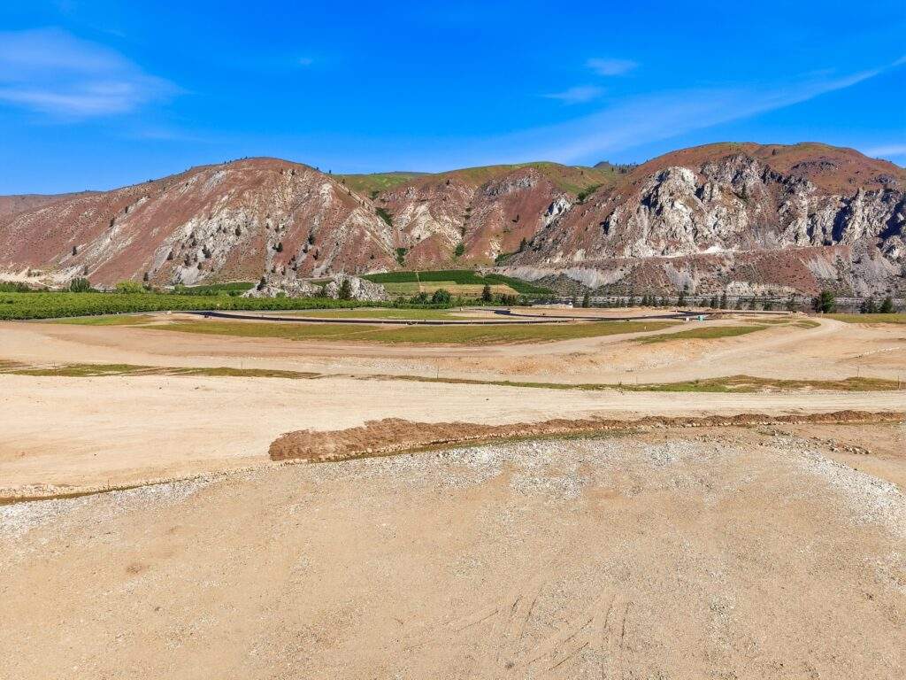 A dry, barren landscape with patches of dirt and gravel, set against hills with sparse vegetation under a clear blue sky, typical of North Central Washington near Lake Chelan.