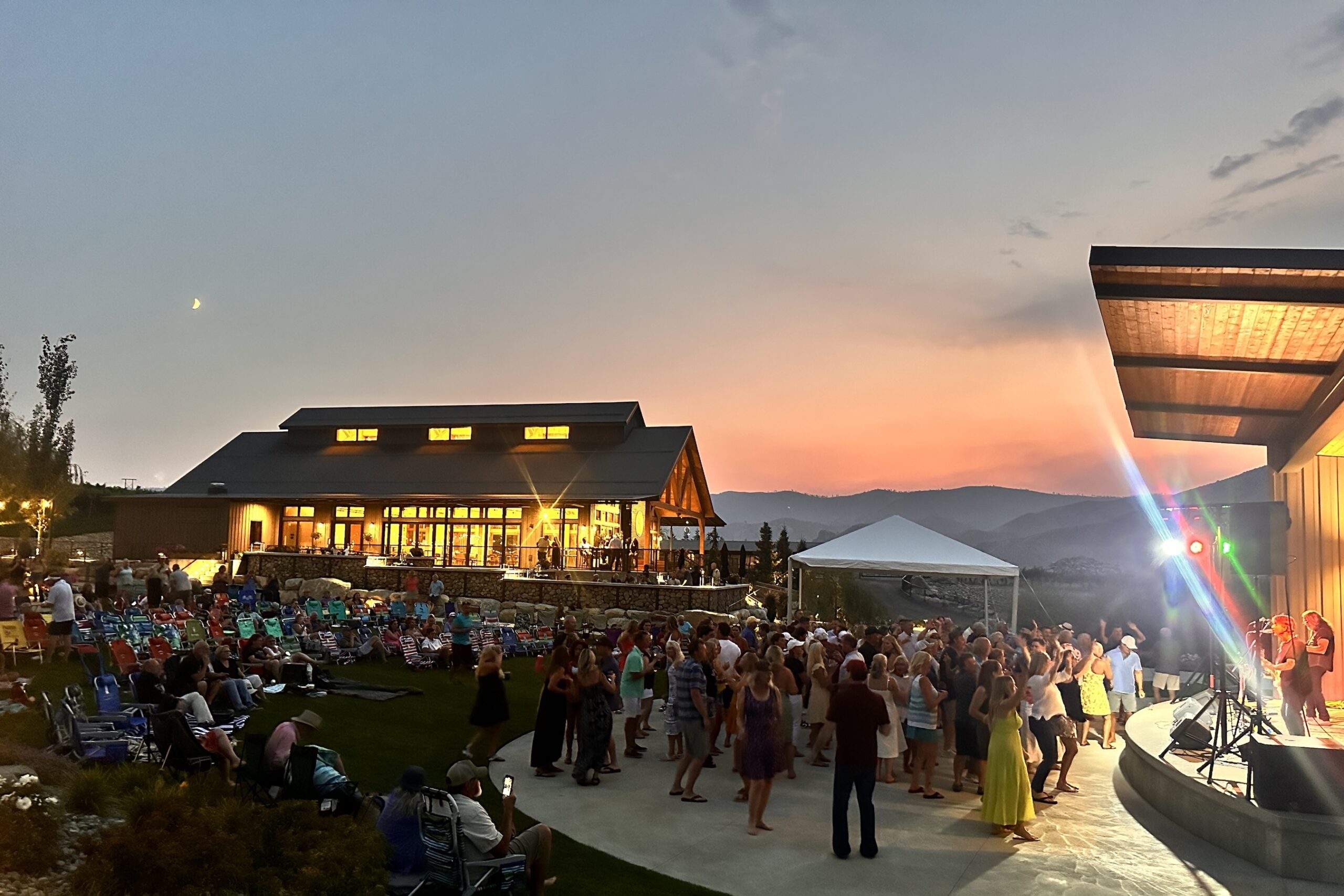 A crowd gathers on a patio near a stage with live musicians at sunset, enjoying vineyard views at Rocky Pond Estate near Lake Chelan, with a modern building and mountains visible in the background.