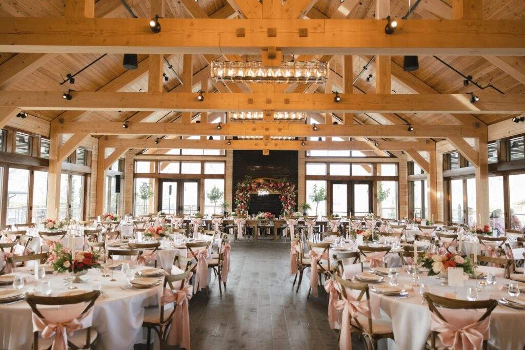 Spacious banquet hall with wooden beams, set up for an event with round tables, floral centerpieces, and pink chair sashes, facing a fireplace with a floral arrangement—enjoy vineyard views near Lake Chelan.