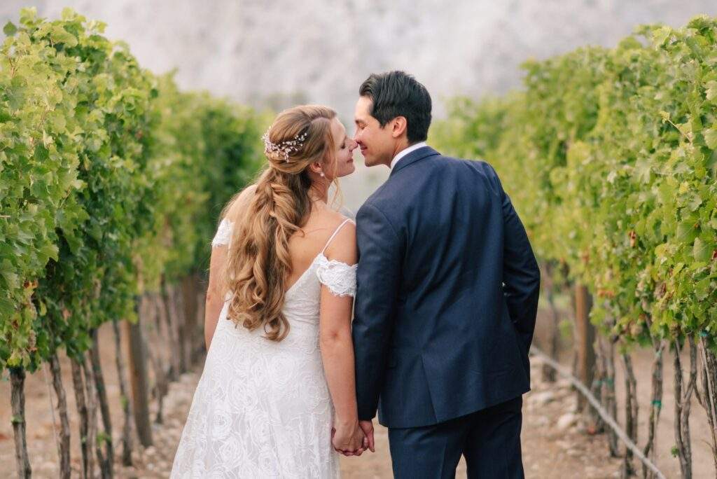 A bride and groom hold hands and touch noses while standing between rows of grapevines, soaking in stunning vineyard views at Rocky Pond Estate in North Central Washington.