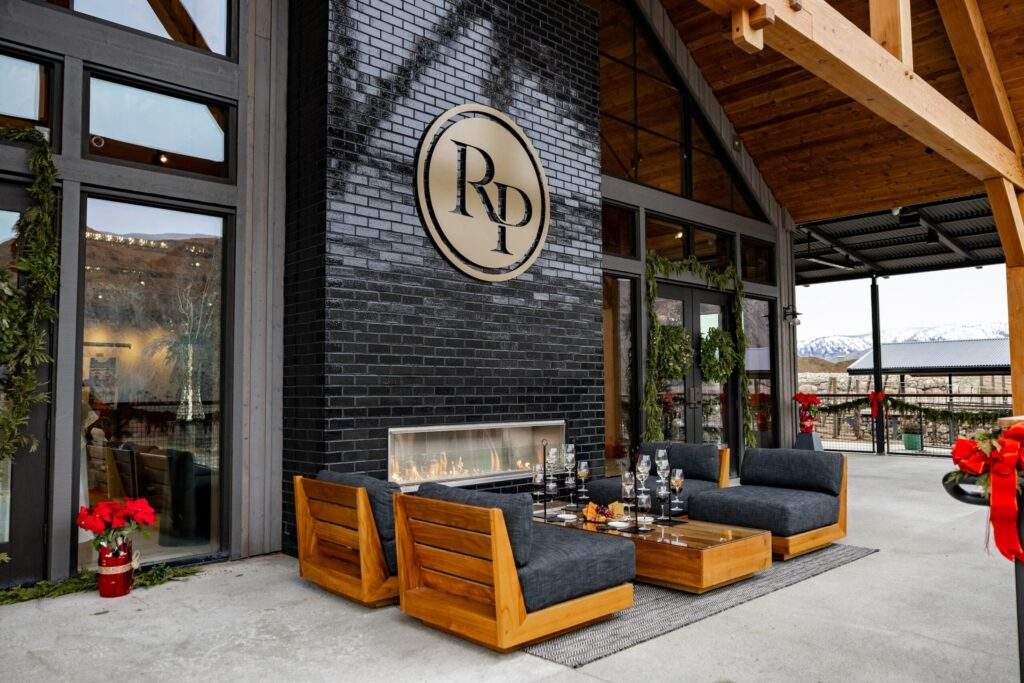 Outdoor patio with modern wooden seating, a low table set with glasses, and a black brick fireplace beneath a large "RD" logo on the Rocky Pond Estate facade, offering scenic vineyard views in North Central Washington.
