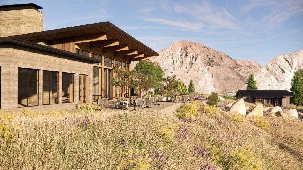 Modern building with large windows and outdoor seating overlooks a field of tall grass and wildflowers, with rocky mountains in the background under a blue sky.