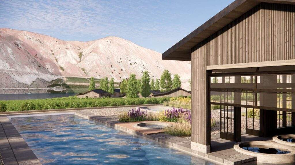 A modern outdoor pool and spa area with wooden buildings, landscaped greenery, and mountains in the background under a blue sky.