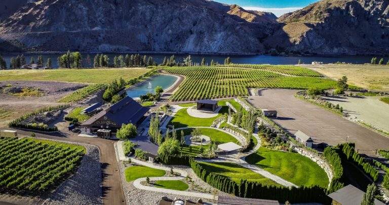 Aerial view of Rocky Pond Estate, a vineyard and winery with landscaped gardens and parking lot, nestled in green fields by a river and mountains near Lake Chelan in North Central Washington.