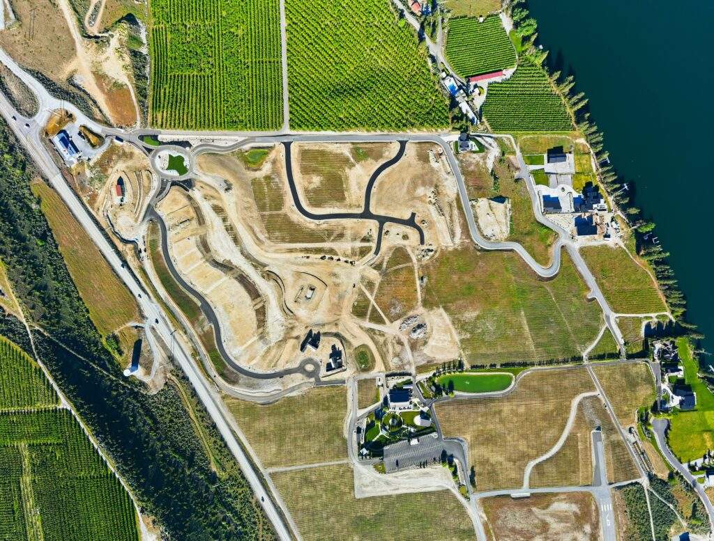 Aerial view of a construction site with winding roads, surrounded by fields, vineyard views from Rocky Pond Estate, and a body of water near Lake Chelan in the upper right corner.