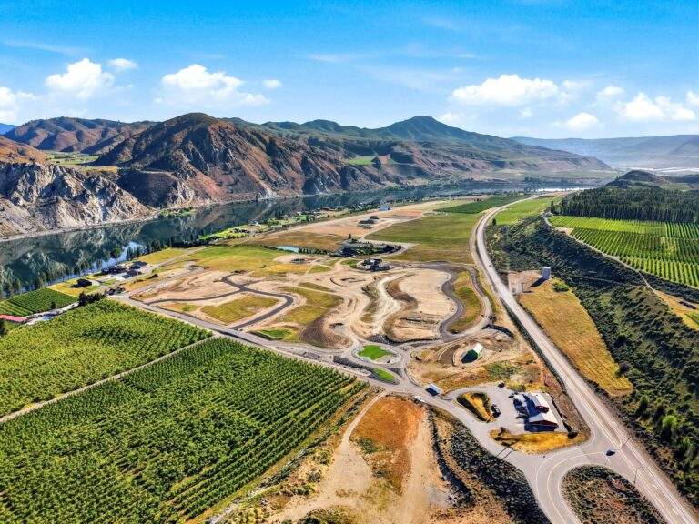 Aerial view of a rural landscape near Lake Chelan, with cultivated fields, winding roads, a river, and mountains in the background under a partly cloudy sky—offering picturesque vineyard views.