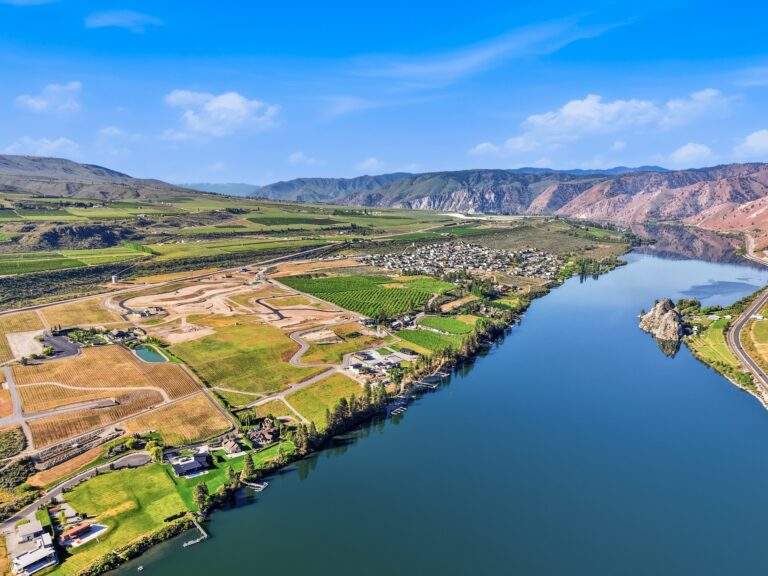 Aerial view of a river bordered by farmland, a small town, roads, and hills under a blue sky with scattered clouds—featuring vineyard views near Lake Chelan and the scenic Rocky Pond Estate.
