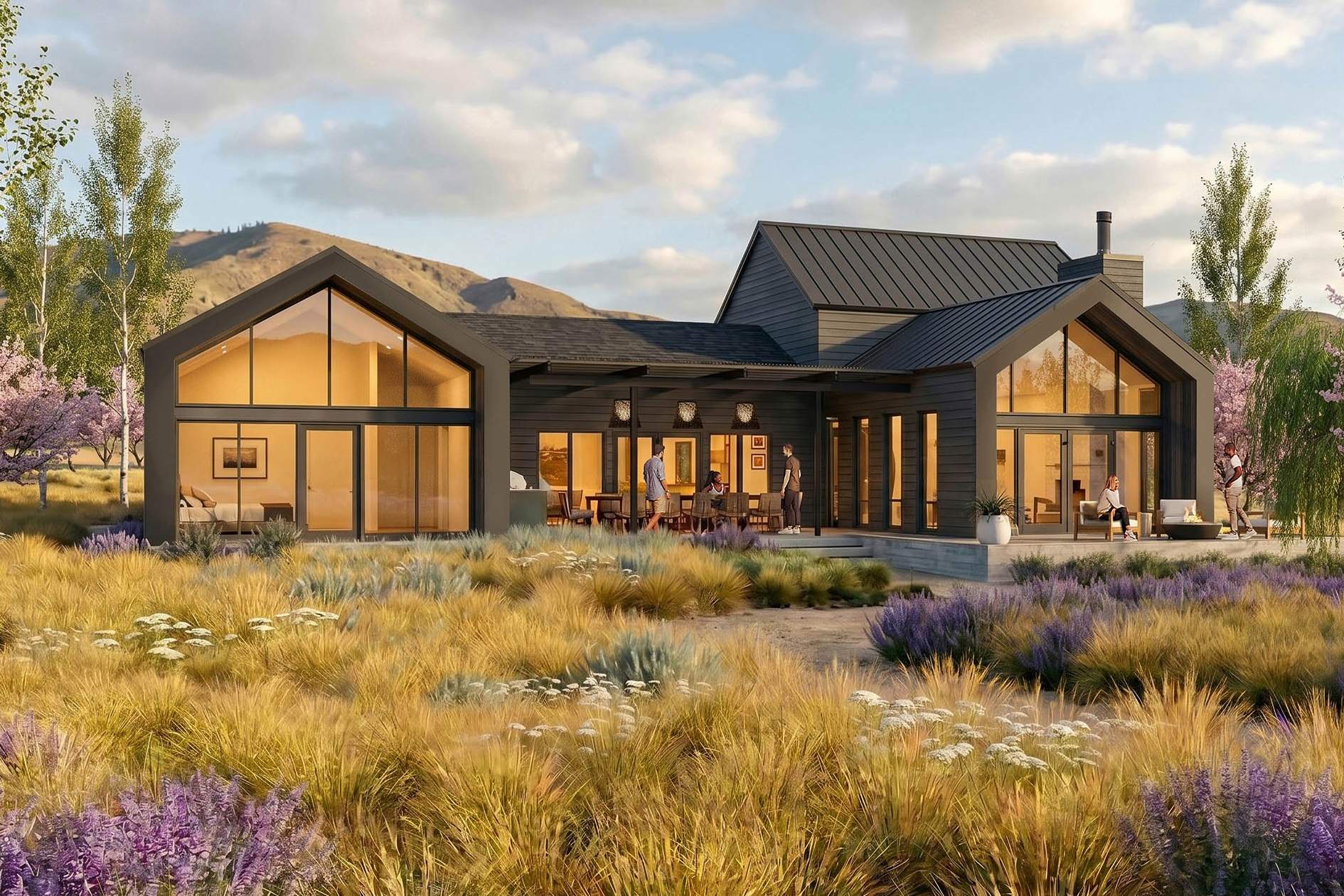 Modern single-story house with large windows and dark exterior, surrounded by grasses and wildflowers, set against a backdrop of hills and partly cloudy sky near Lake Chelan in North Central Washington.